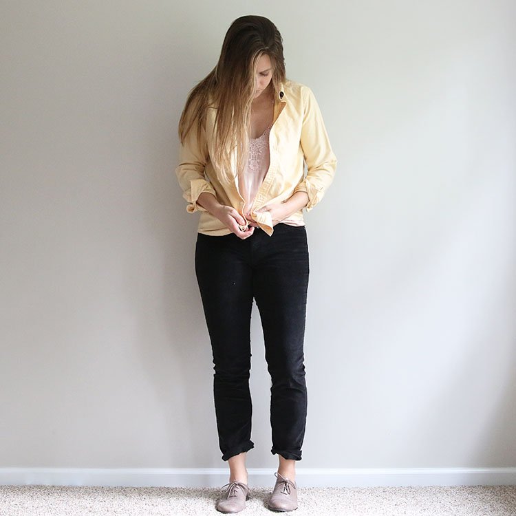 woman tying a knot in a yellow shirt over a pink cami and black corduroy pants