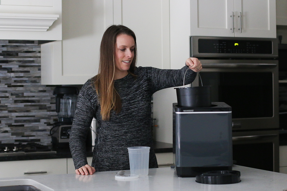 Carbon Emission Analysis Of The Vitamix FoodCycler: Woman holding Food Cycler bucket in the Food Cycler appliance sitting on the kitchen counter. 